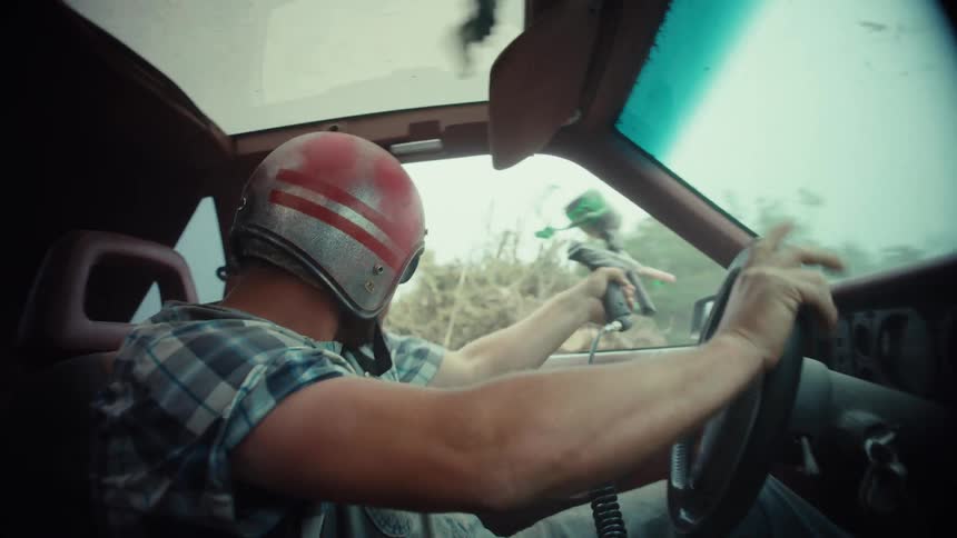 Slow motion shot from inside a vintage car shows a man in a helmet driving and shooting a paintball gun