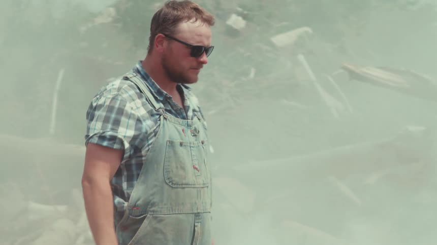 A frustrated man in overalls stands by his smoking, broken-down car in a junkyard before kicking a ball and walking away