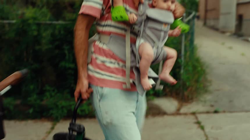 A busy father multitasks on a walk, carrying his baby in a front carrier and his toddler on his shoulders while pushing a stroller and holding a small bike