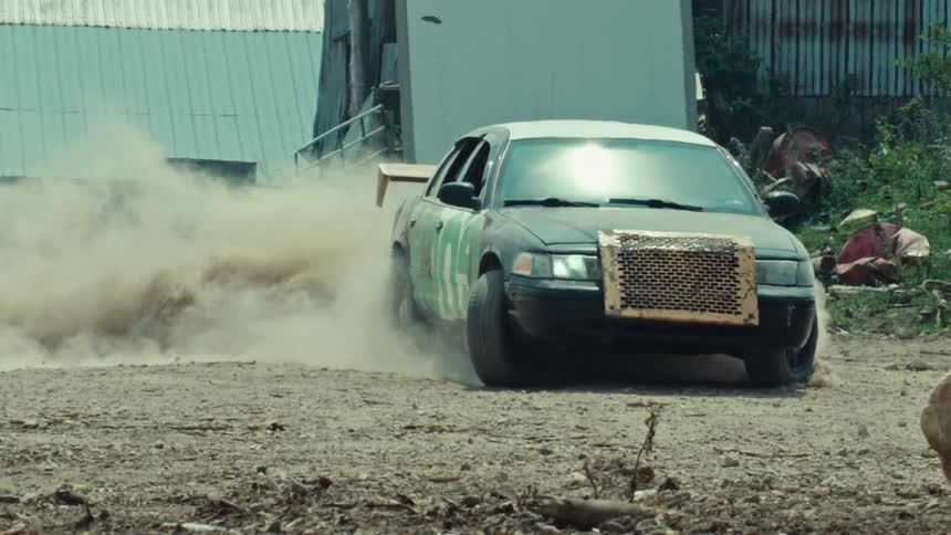A custom black and white sedan with green lettering does a powerful burnout on a dirt lot, kicking up a massive cloud of dust in dramatic slow motion