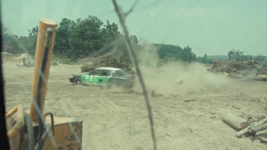 Slow motion view from inside heavy machinery as a beat-up car does donuts, kicking up a massive cloud of dust in an industrial lot
