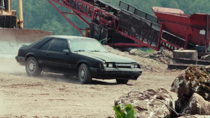 Two cars race and drift in slow motion on a dirt track in a dusty, industrial quarry