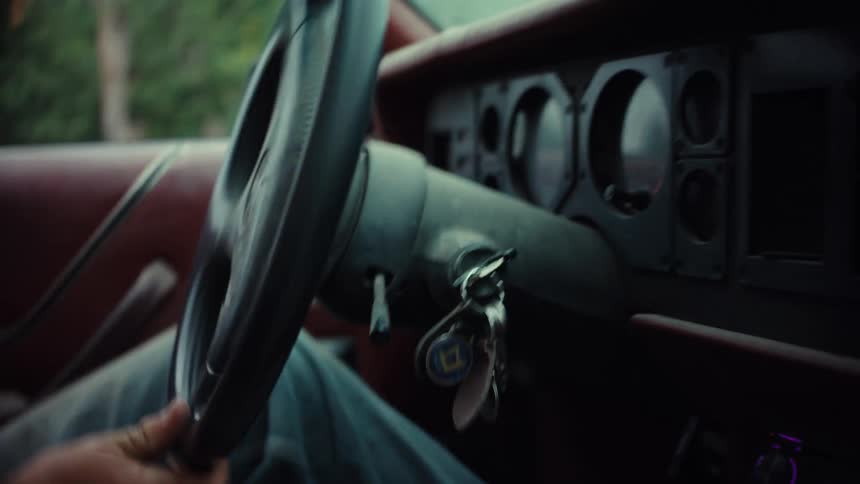 A gritty, shaky handheld shot from inside a vintage car focuses on the dashboard and steering wheel as a person drives