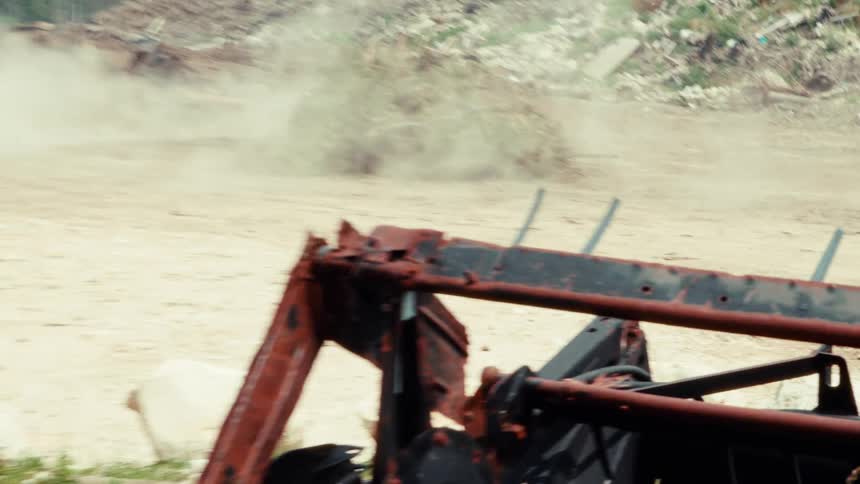A demolition derby car with "GUCK" spray-painted on the side drives through a dusty junkyard, kicking up dirt and crashing into a pile of brush in slow motion
