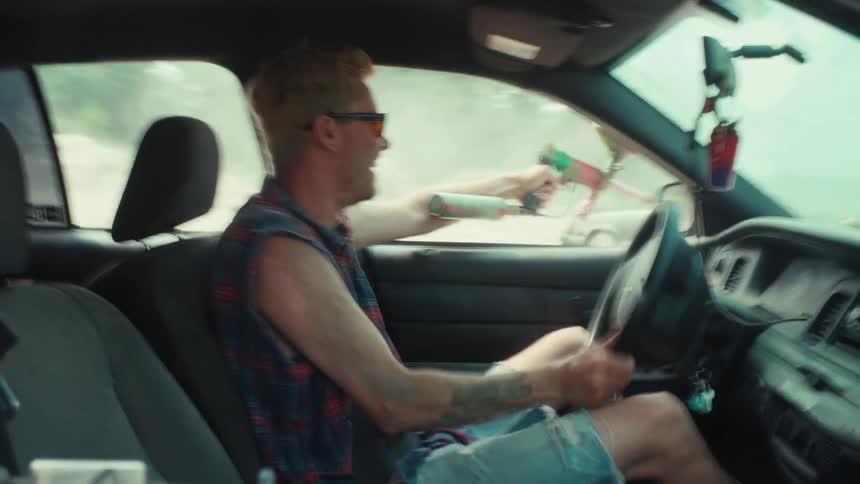 A smiling man with blonde hair enjoys a reckless car ride while shooting a paintball gun out the window in slow motion