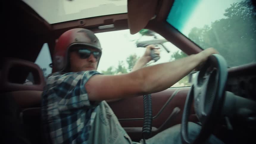 A man wearing a helmet and sunglasses chaotically drives a car while shooting a paintball gun out the window