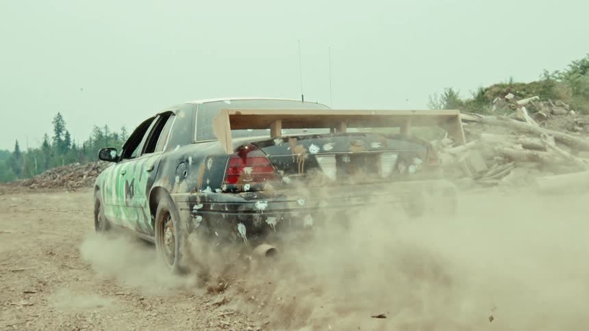 A modified race car with a large wooden spoiler does a burnout in super slow motion, kicking up a large cloud of dust on a dirt road