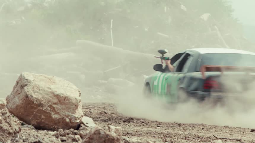 A customized sedan speeds down a dusty dirt road while a passenger leans out the window and aims a paintball gun in slow motion