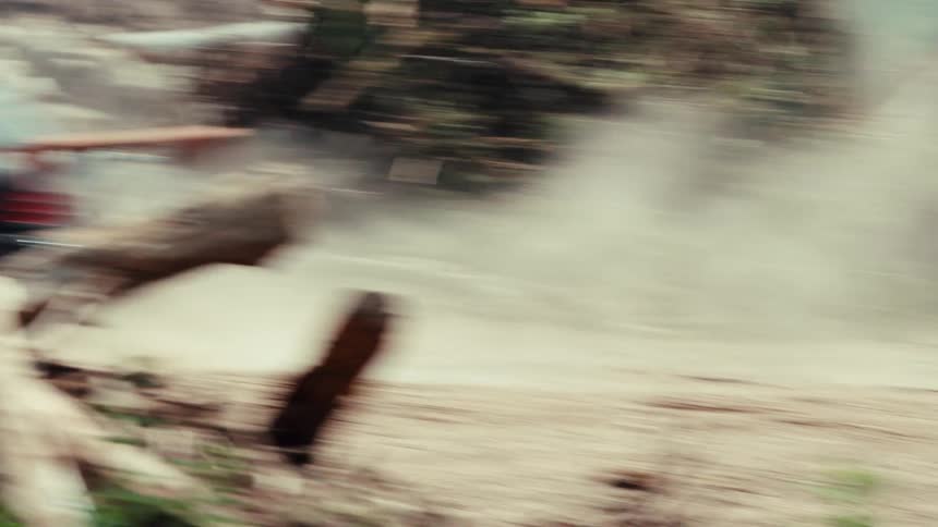 Slow motion tracking shot of two modified cars racing on a dirt track while passengers lean out and shoot paintball guns at each other