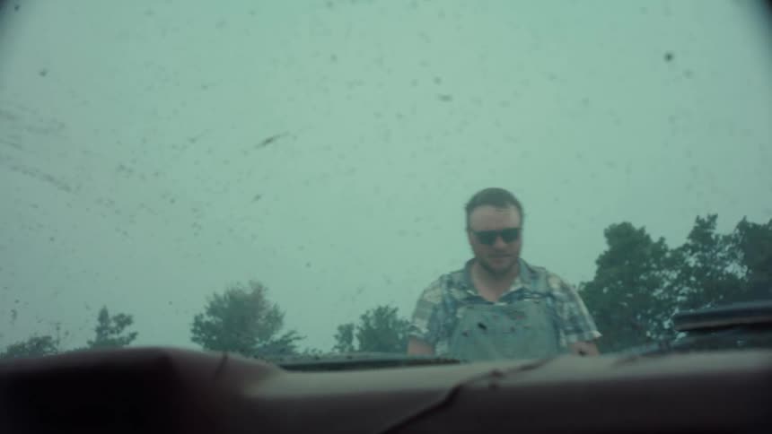 Furious man in overalls seen through a dirty windshield yelling and slamming his hands down on the hood of a car in slow motion