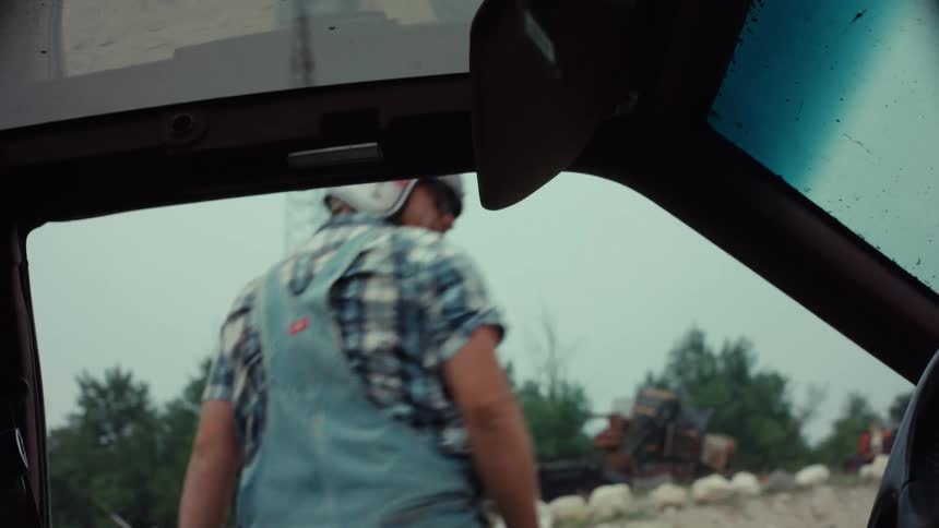 A man in overalls and a helmet climbs out of the window of his car at a rural junkyard