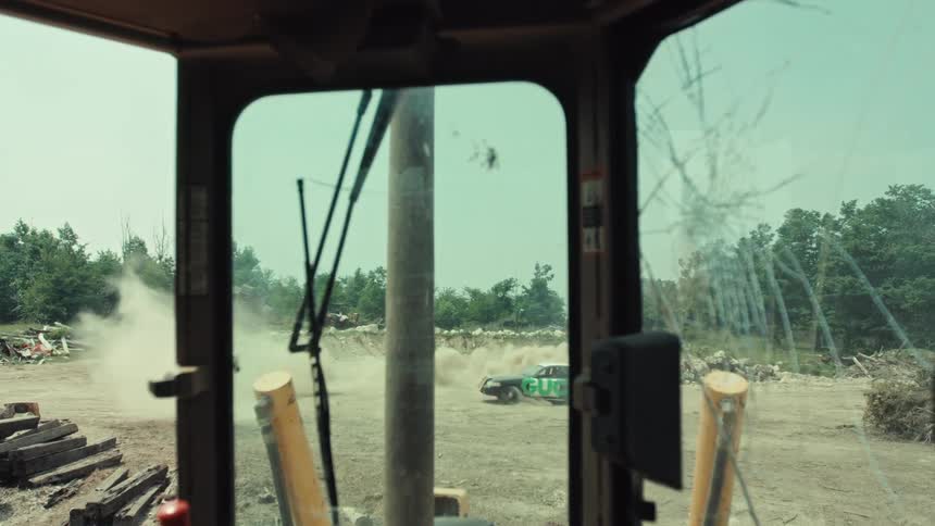 Point of view from a bulldozer cab with a cracked windshield watches a derby car spin out and crash in a huge cloud of dust