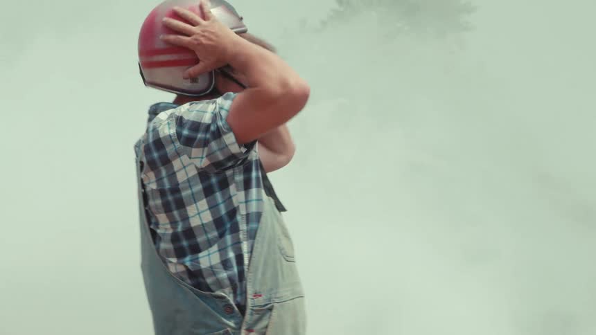 A frustrated man in overalls and a helmet throws his helmet on the ground next to a smoking car in a junk yard