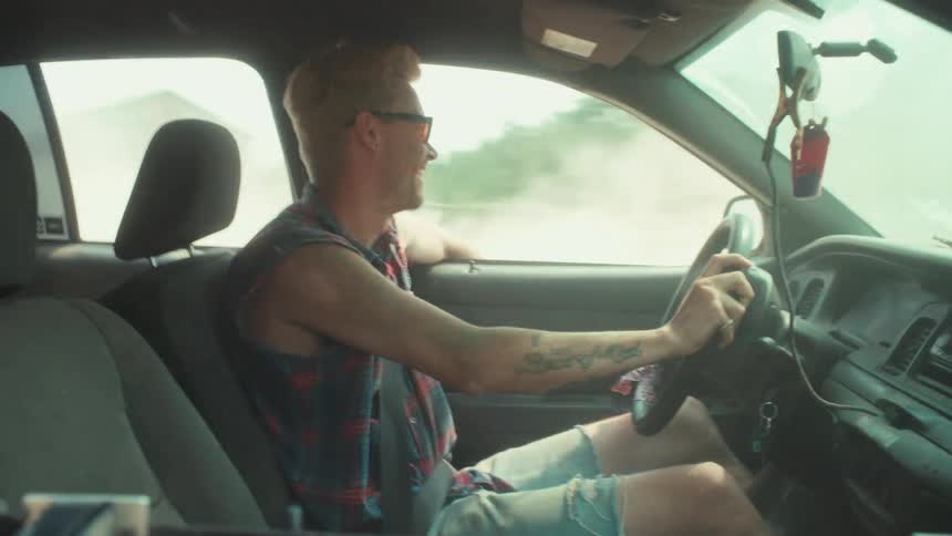 A carefree young man with sunglasses and a cutoff plaid shirt smiles and enjoys a bumpy ride while driving down a dusty road