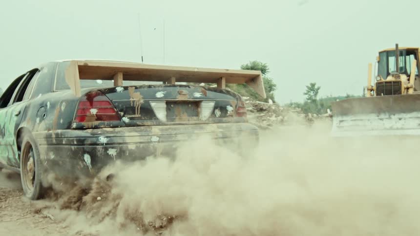 A custom black race car with a large wooden spoiler does a burnout in a dusty junkyard in slow motion