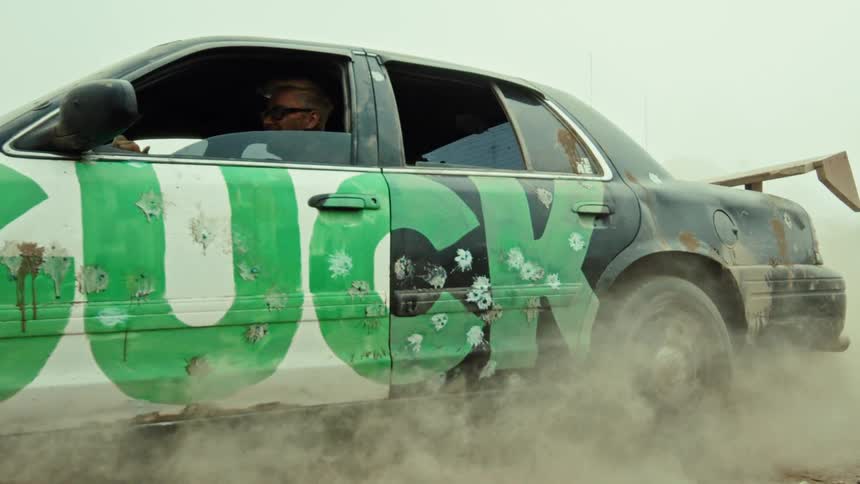A modified black derby car with 'LUCKY' spray-painted on its side performs a burnout in slow motion, kicking up a massive cloud of dust