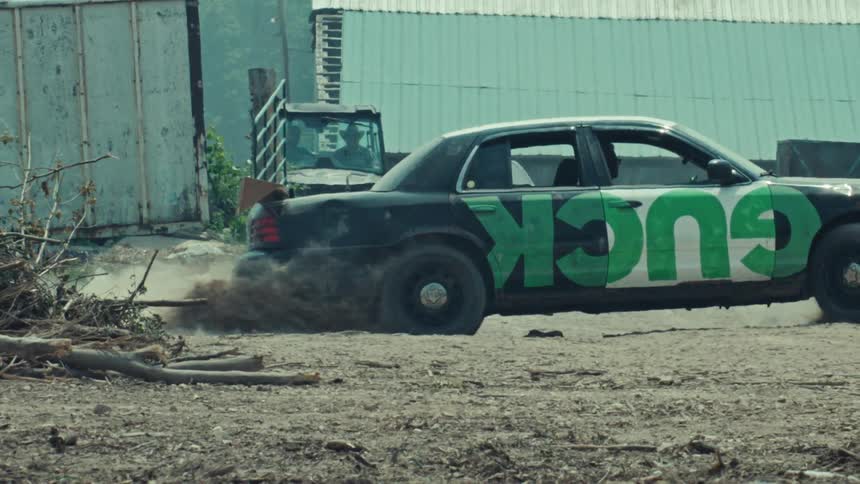 In slow motion, a black sedan with 'GUCK' graffiti on its side performs a powerful burnout, spewing dust and smoke from its spinning tire