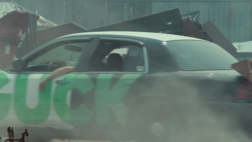 A modified black and white car with a homemade spoiler and a large green logo kicks up dust as it drives in slow motion