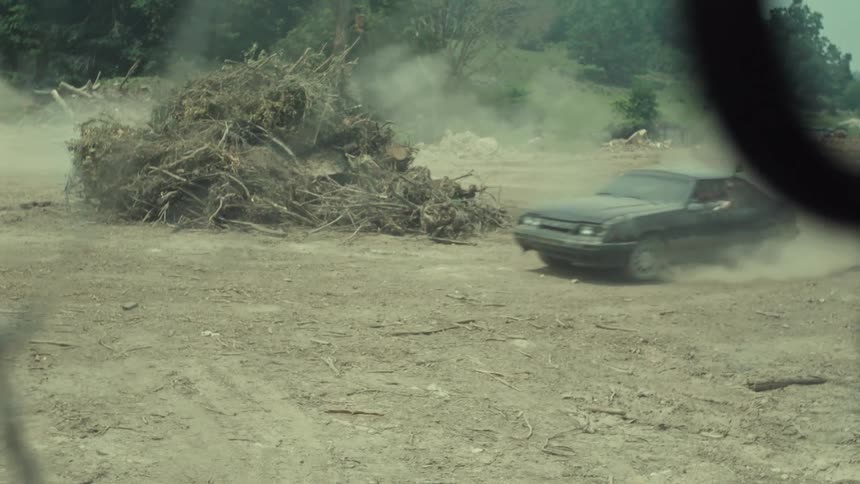 A man in a vintage black Mustang does donuts in a dusty lot while holding a paintball gun out the window