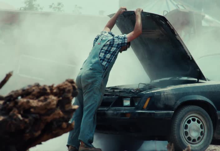 A frustrated man in overalls angrily gets out of his smoking muscle car, kicks it, and gives up after the engine breaks down