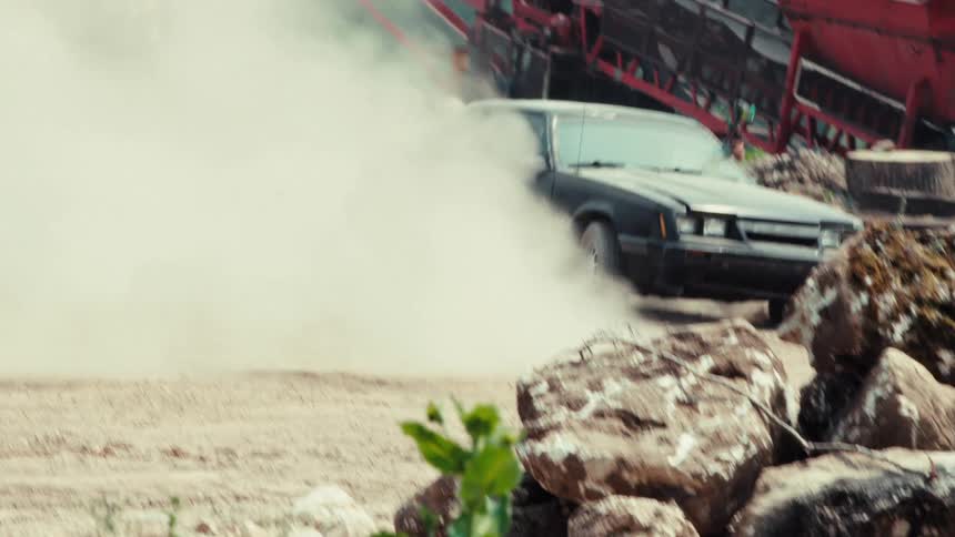 Two cars race through a dusty industrial lot while passengers shoot paintball guns at each other in an intense action sequence