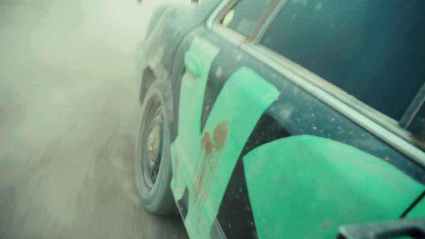 Seamless loop of a black and green demolition derby car speeding down a dirt road, kicking up dust in slow motion
