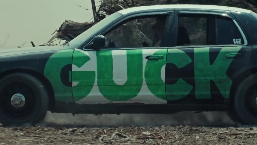 A customized black and white sedan with 'GUCK' in green letters does a powerful burnout on a dirt lot in slow motion