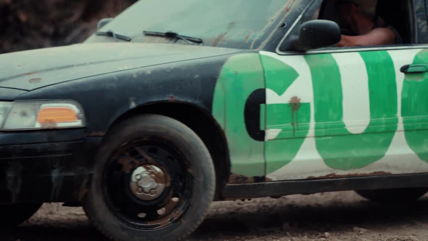 A beat-up derby car with 'GUCK' painted on the side drives through a post-apocalyptic junkyard setting