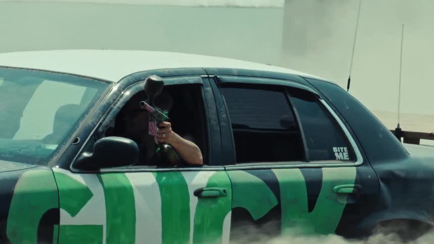 Slow motion shot of a man aiming a paintball gun from the window of a car doing a smoky burnout