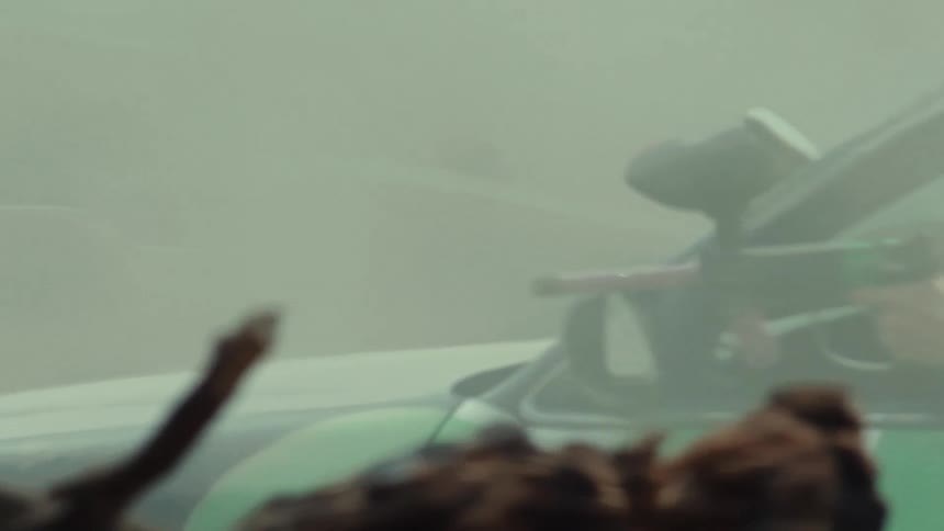 A person aims a paintball gun out of a car window amidst thick smoke in slow motion