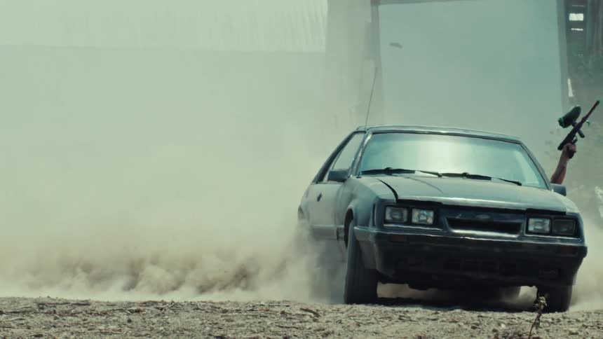 A black muscle car kicks up a cloud of dust doing a burnout while a man with a paintball gun leans out the window in slow motion