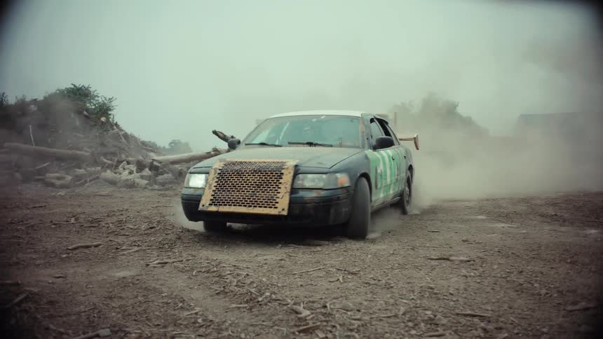 A souped-up demolition derby car drives in slow motion through a dusty, apocalyptic junkyard