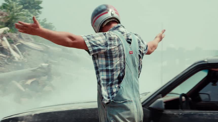 A man in a helmet and overalls gets out of his broken-down classic car, which is billowing smoke, and opens the hood in slow motion
