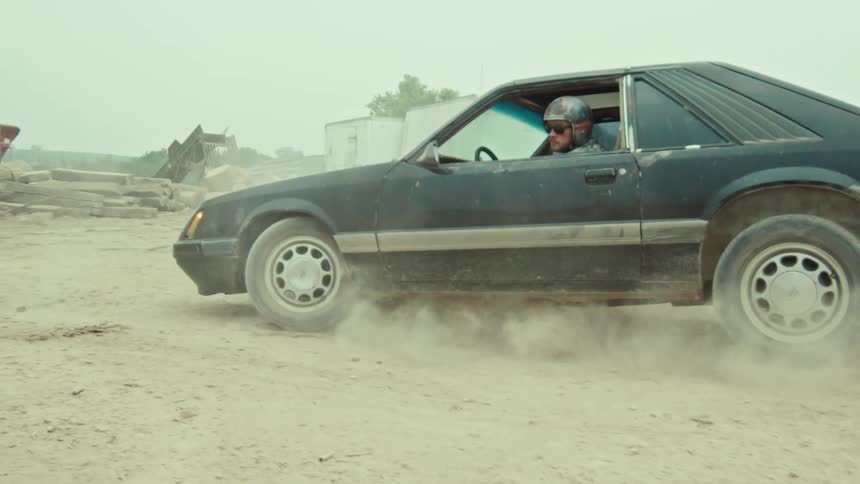 A man in a helmet does a burnout in a vintage black muscle car on a dirt road, kicking up a cloud of dust in slow motion
