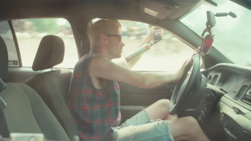 A rebellious man in a sleeveless plaid shirt drives a car while joyfully shooting a paintball gun out the window in slow motion