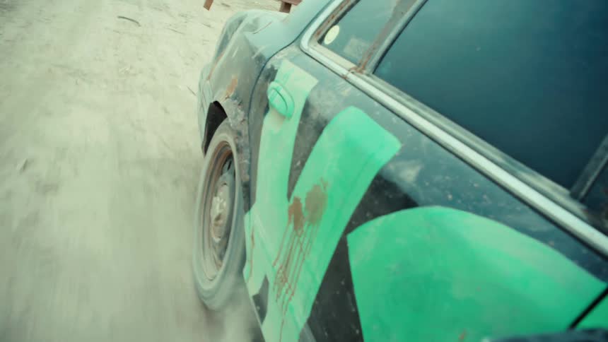 A close-up slow-motion shot shows a demolition derby car's wheel spinning in place, kicking up a large cloud of dust from the dirt track