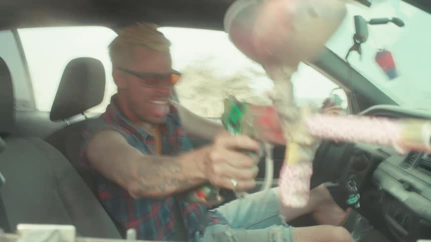 A wild man with blonde hair drives erratically while shooting a paintball gun out of his car window in slow motion