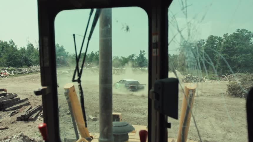 From the cracked windshield of a bulldozer, a green and black stock car drifts through a dirt lot kicking up a huge cloud of dust