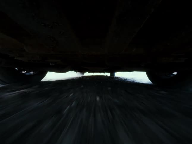 A unique low-angle POV shot from underneath a car as it speeds down a rough, dark road and then comes to a stop