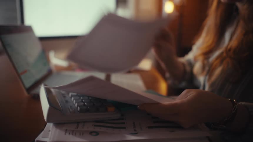 A focused woman with red hair and glasses diligently goes through financial reports and paperwork at her home office desk