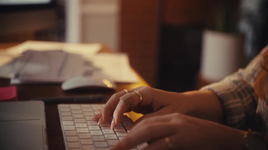 A woman's hands type on a computer keyboard in slow motion at her desk in a warmly lit room