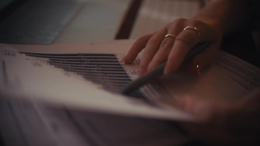 A person's hands trace the data on a printed financial chart with a pen while working at a dimly lit desk with a laptop in the background