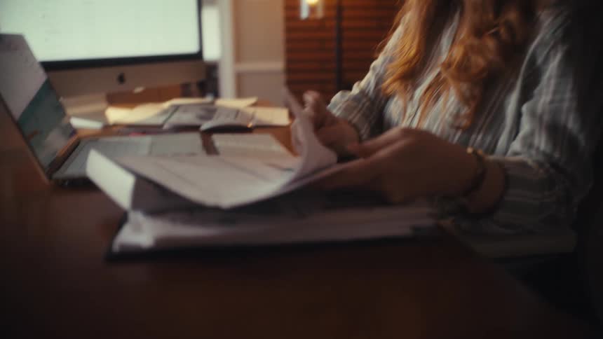 A focused woman reviews paperwork and financial reports while working late into the night at her home office desk