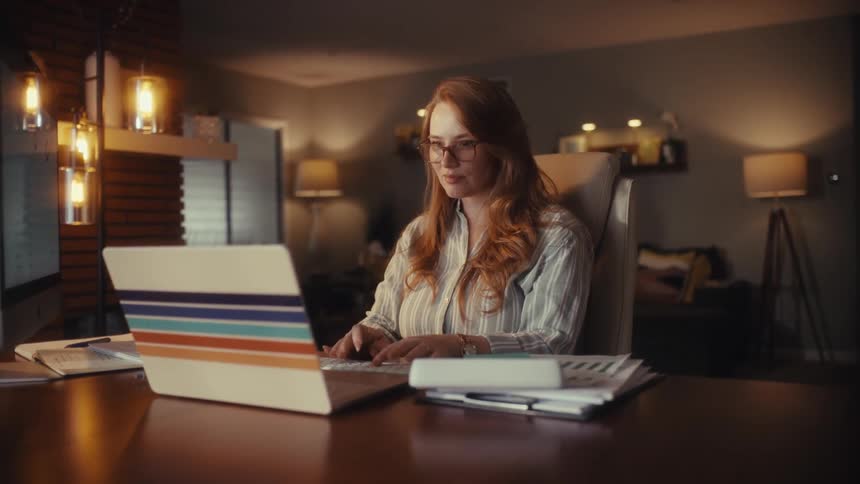 A focused woman with red hair and glasses works late at night in her home office, typing on her laptop