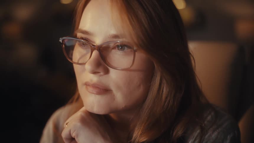 A focused redhead woman with glasses works late into the night on her laptop in a dimly lit home office