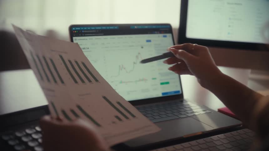 A financial analyst compares data from a printed report with a live stock market chart on a laptop screen