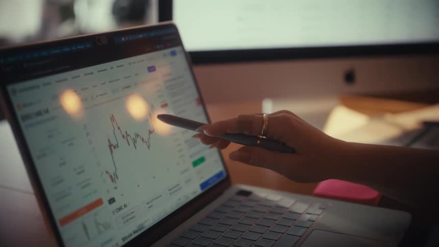 A business person reviews financial data on printed charts before transitioning to analyze market trends on a laptop and desktop computer