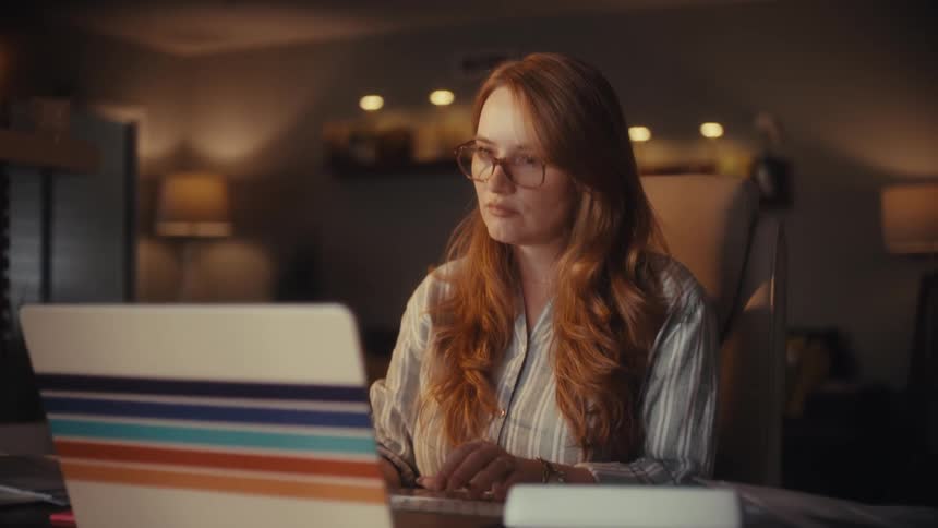A thoughtful woman with red hair and glasses works late into the night on her laptop, pausing to think in her dimly lit home office