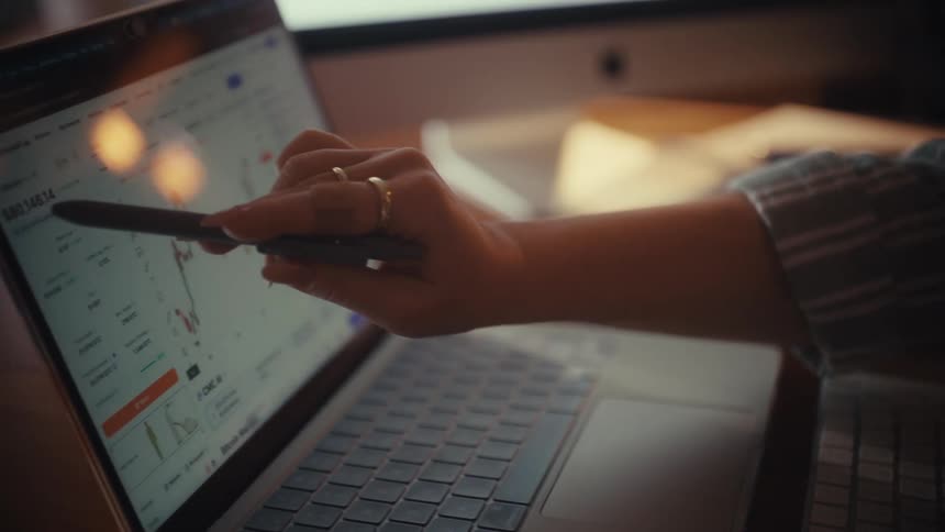 A person reviews financial portfolio documents before using a stylus to analyze stock market data on a laptop