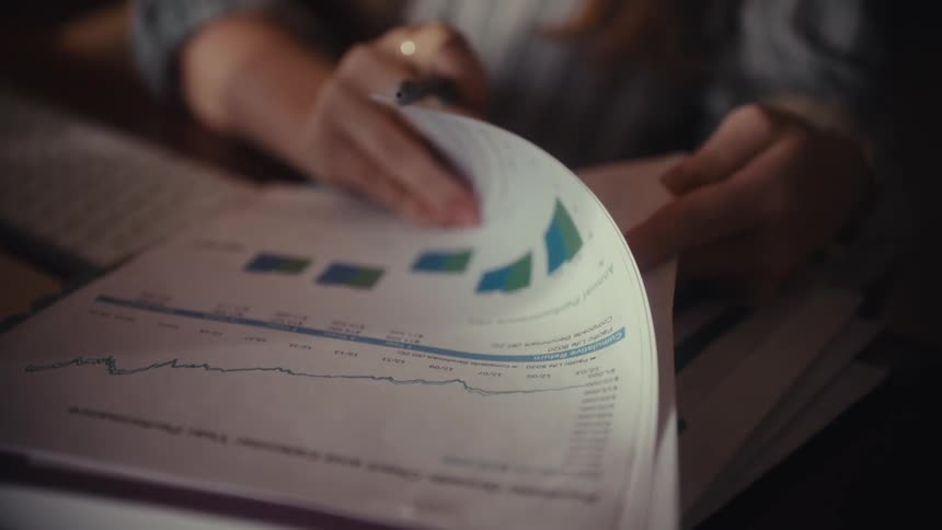 A business analyst slowly reviews portfolio growth charts and performance data on paper documents